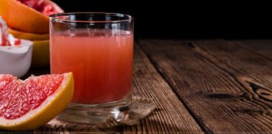 A glass of grapefruit juice on a dark brown table, with slices of grapefruit in front of it.