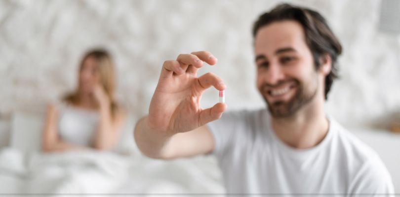 A man sitting in bed with a woman in the background. He is holding up a pill and smiling.