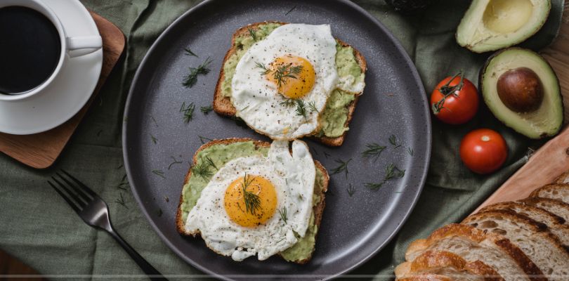 A plate of over easy eggs on top of toast, with bread, coffee and avocado arranged around it on a table.