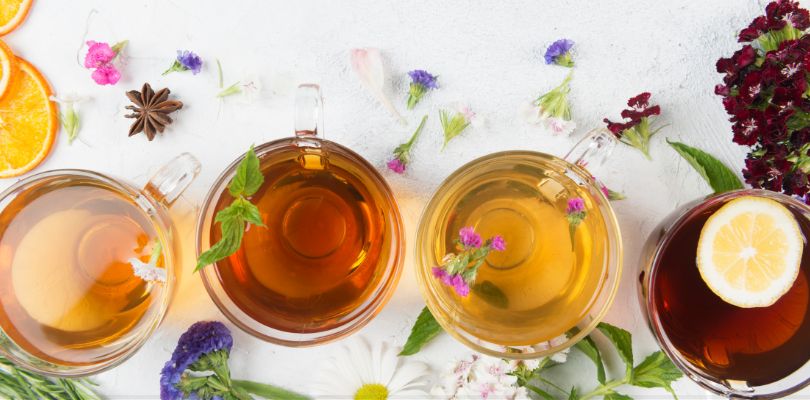 Four clear mugs of different types of tea on a white table, with tea leaves and flowers scattered around them.