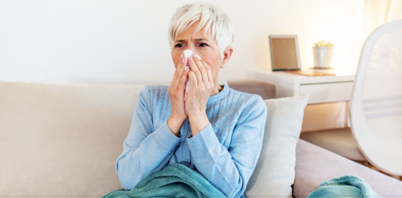 A woman sitting on her couch with a blanket, blowing her nose.